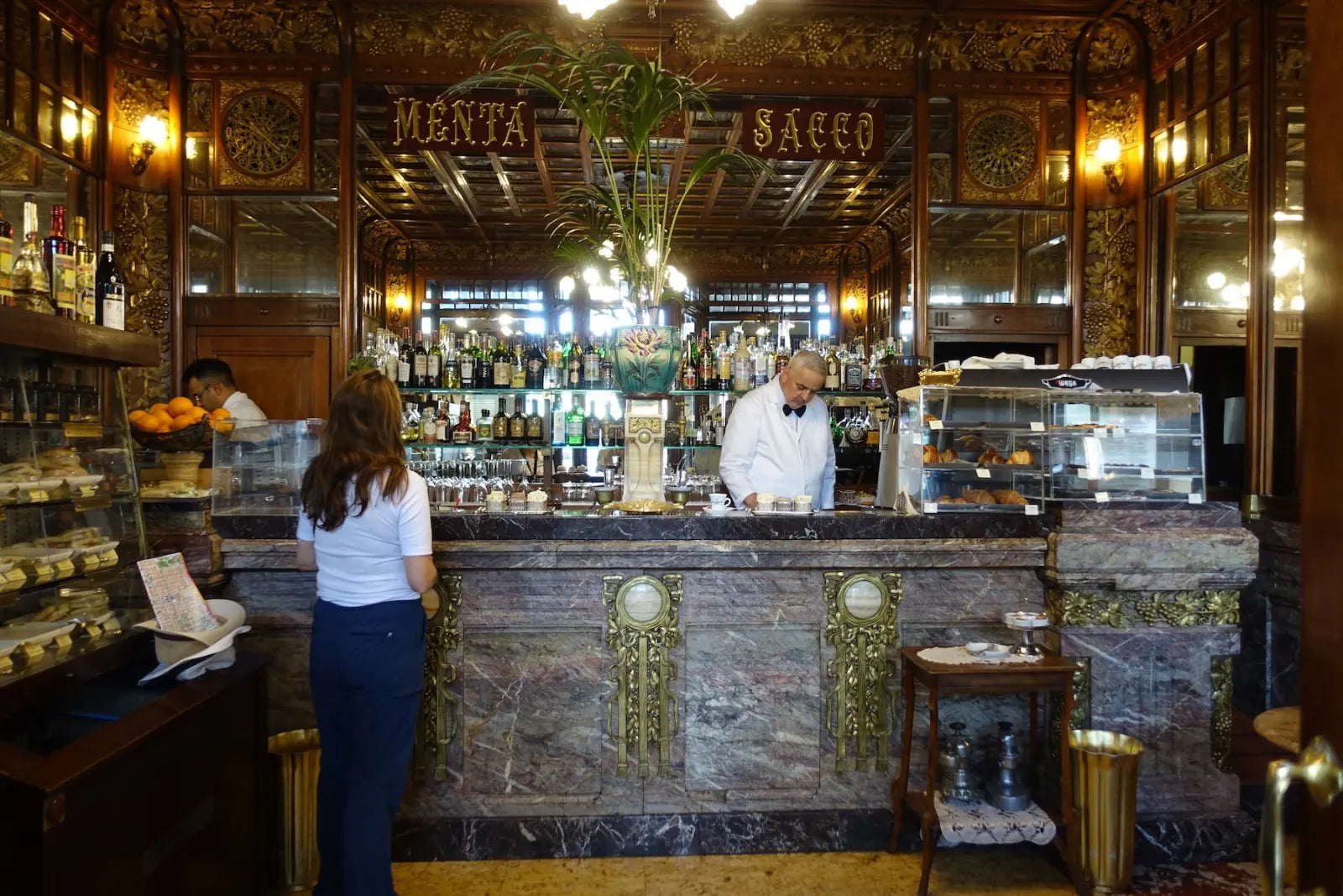 An ornate café counter with a bartender, a customer and pastry cases.