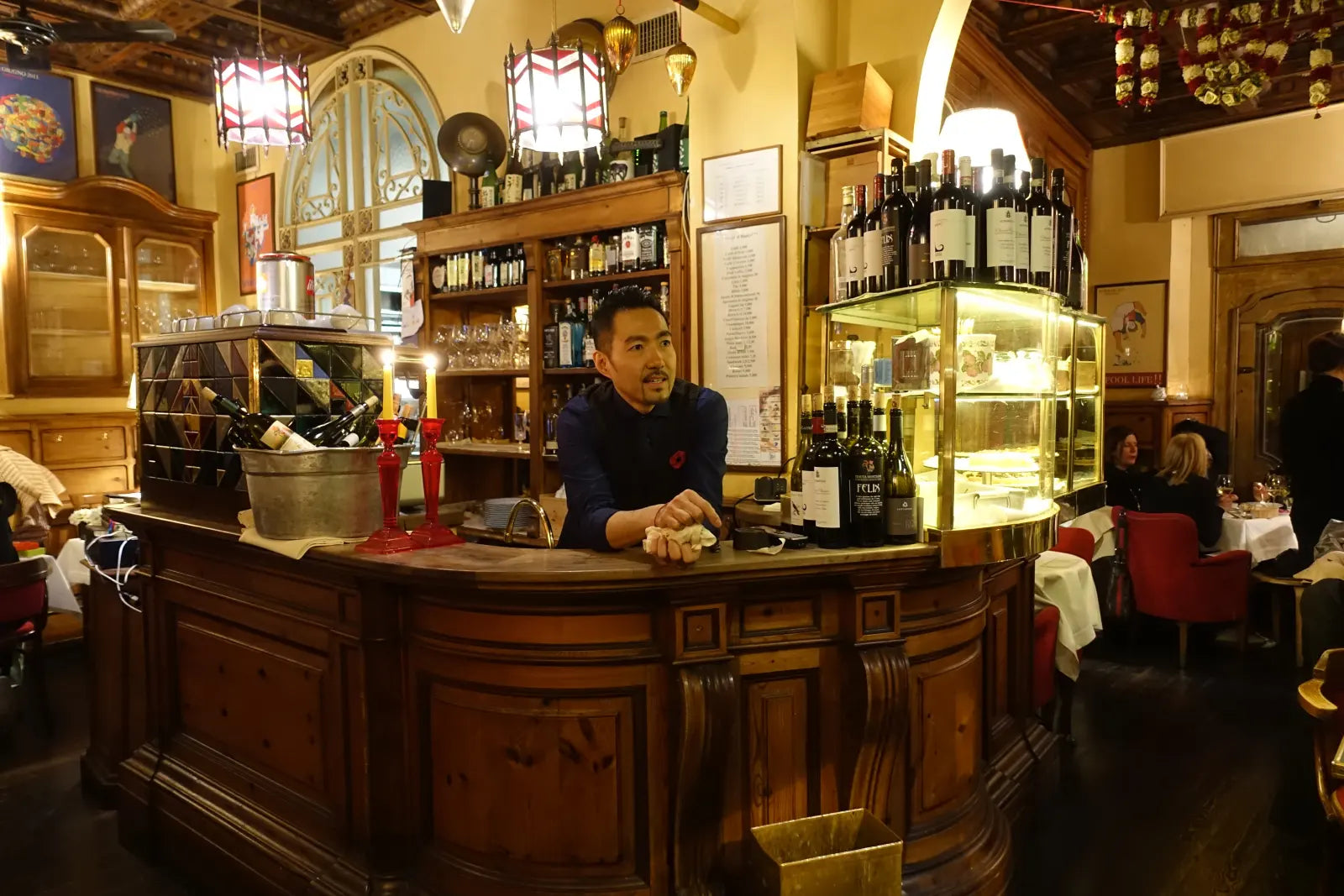 Yusuke Nomiya leaning behind a wooden bar counter with wine bottles beside him.