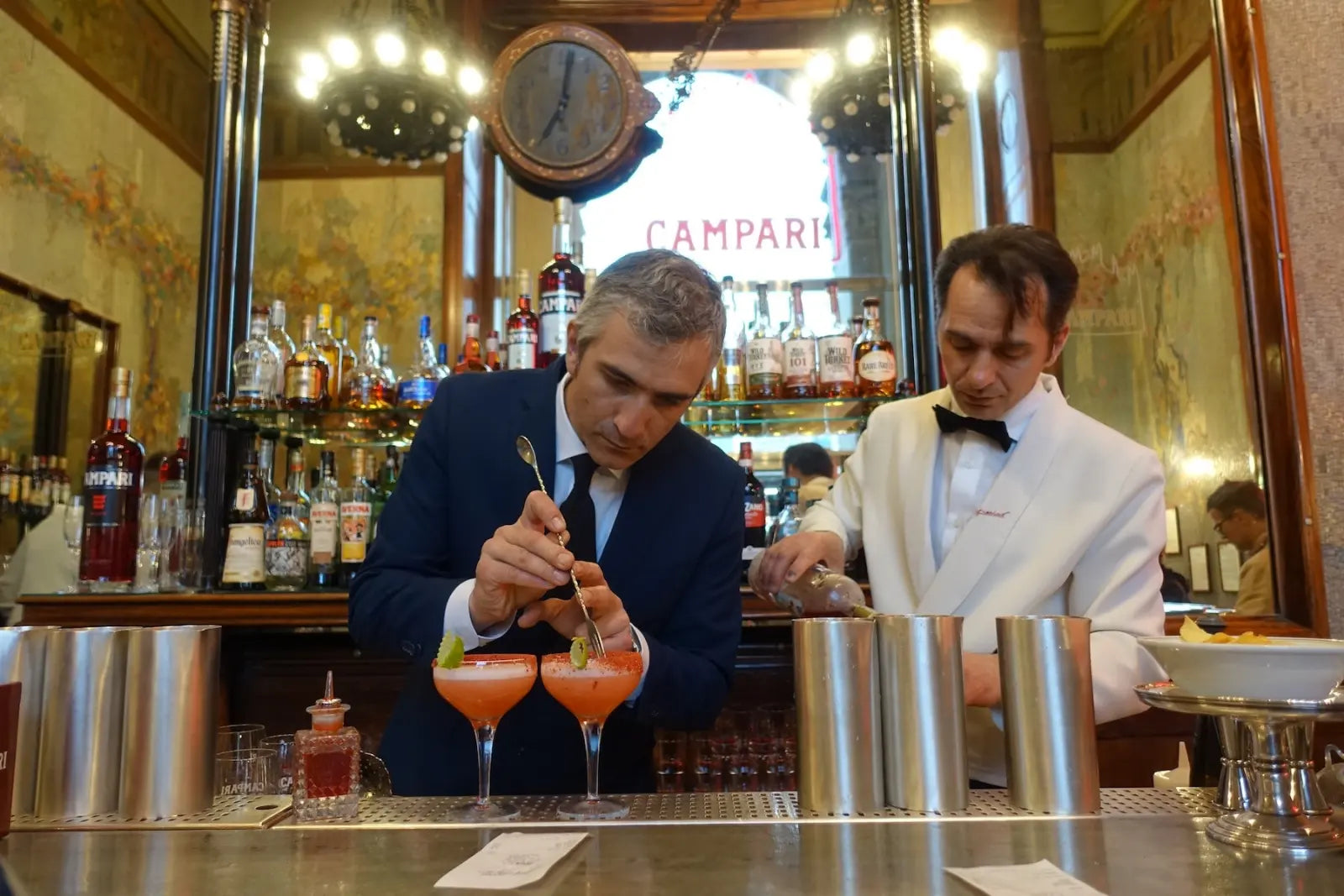 Two men preparing orange-red cocktails behind a bar counter.