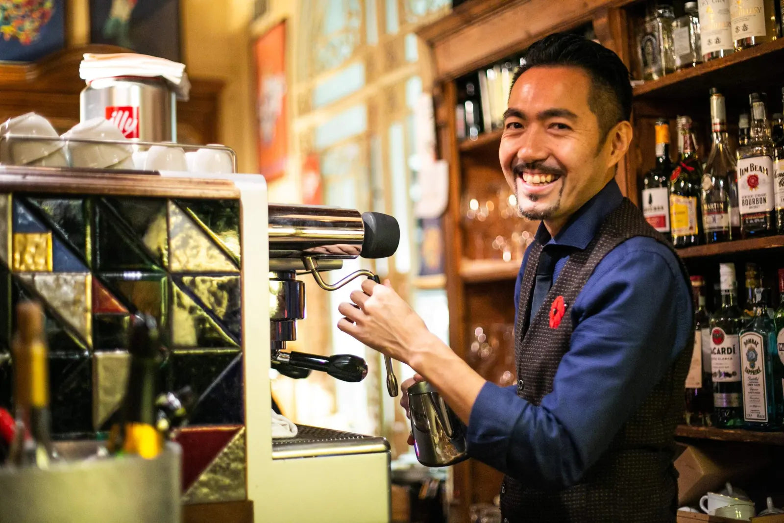 Yusuke Nomiya smiling while holding a metal milk jug beside an espresso machine.