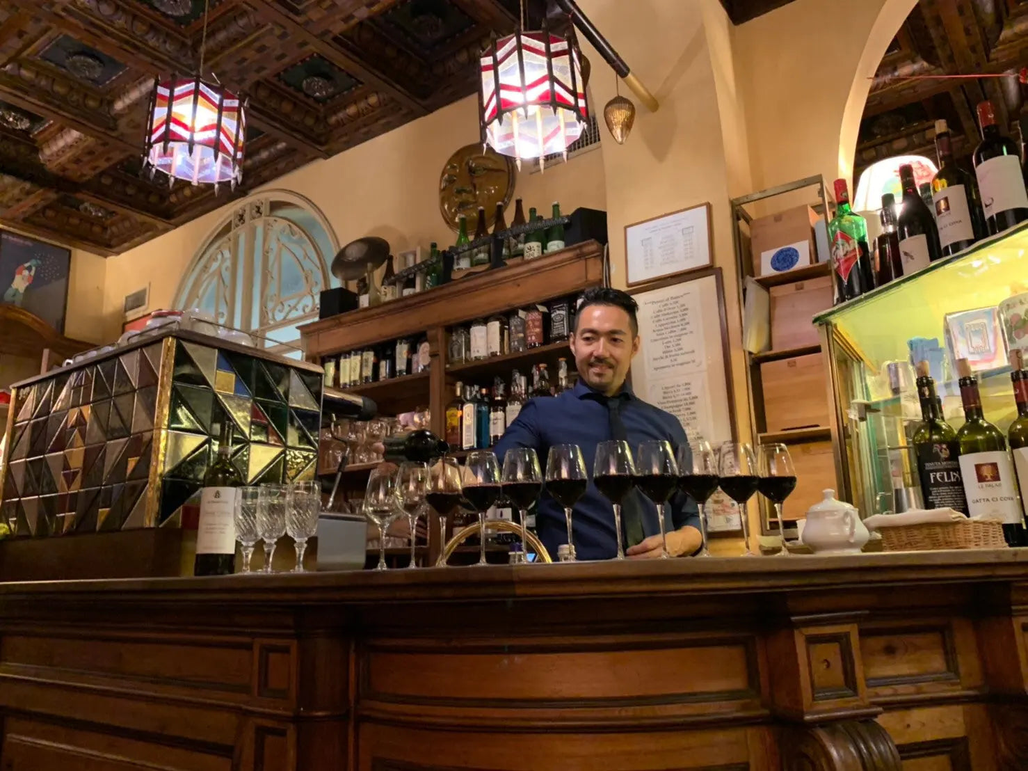 Yusuke Nomiya behind a bar counter with a row of glasses of red wine.